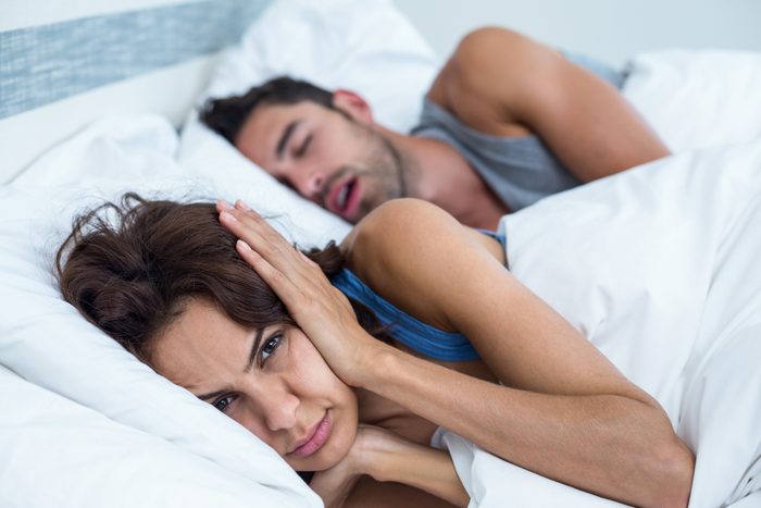 Woman blocking ears with hands while man snoring on bed