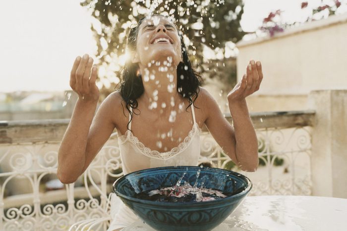 Woman on a Balcony Sits With Her Eyes Close, Splashing Water From a Bowl