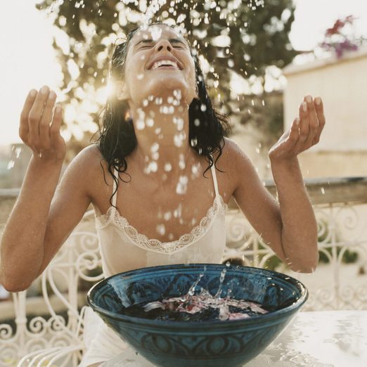 Woman on a Balcony Sits With Her Eyes Close, Splashing Water From a Bowl