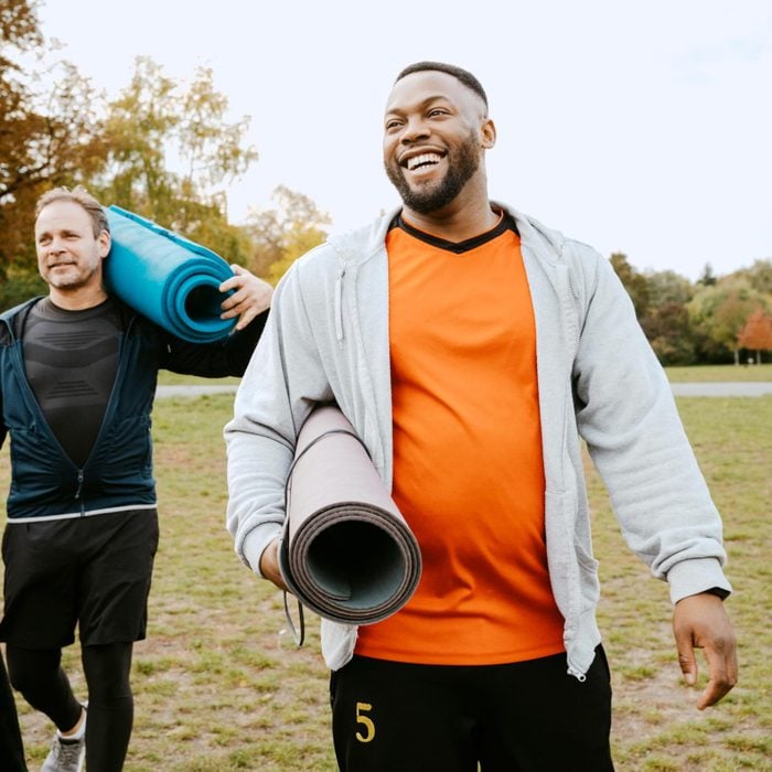 Cheerful man with exercise mat walking by male friend in park