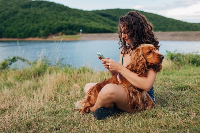 A cocker spaniel dog is in the arms of its owner, who is sitting on the shore of a lake and typing a text message