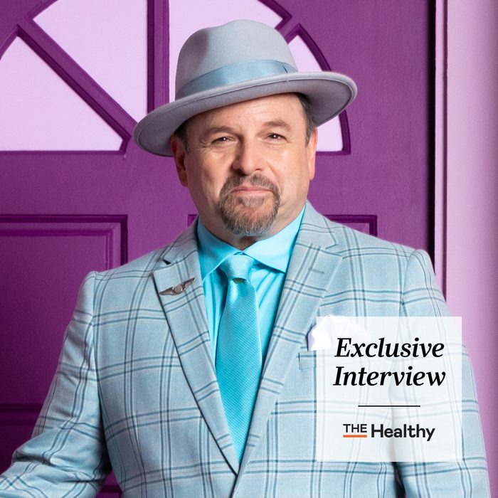 portrait of Jason Alexander in a grey suit and hat with a purple door behind him. The healthy Exclusive Interview stamp is in the borrow right corner.
