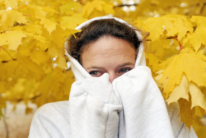 Woman wearing hooded sweatshirt pulled over her face with yellow autumn leaves behind her