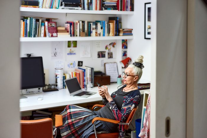 stylish Senior woman sitting in home office with bookshelves and papers hanging on the wall