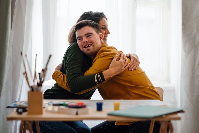 Young man with Down syndrome sitting and embracing with his tutor after painting indoors at home, homeschooling.