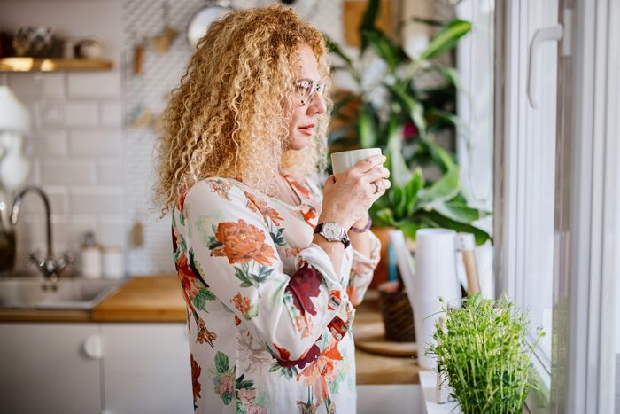A mature woman enjoys a cup of tea and looks out the window from her kitchen