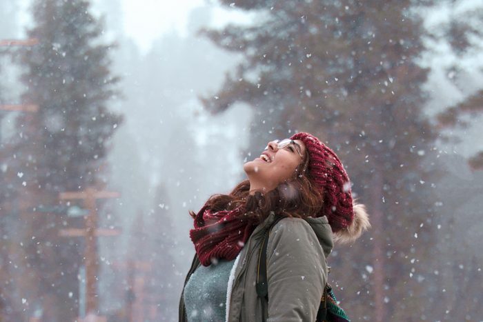 A young woman wearing a hat, coat and scarf opening her mouth the catch the falling snowflakes outside in the winter