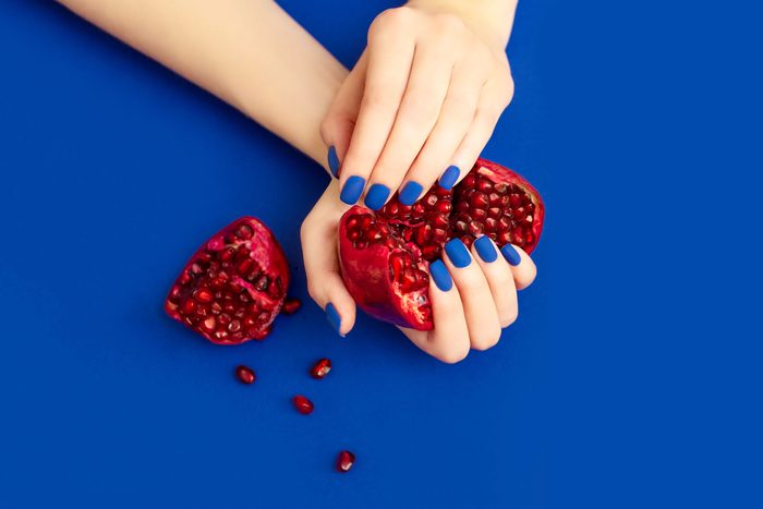woman hands with blue finger nails holding an open pomegranate on a blue background that matches her nails