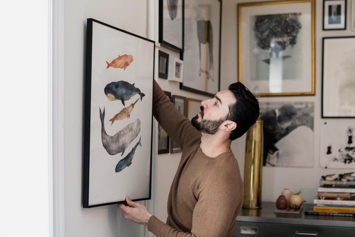 Young man hanging watercolor painting with whales on wall at home
