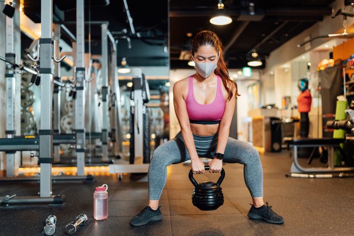 Female athlete wearing protective face mask and lifting kettlebell in gym