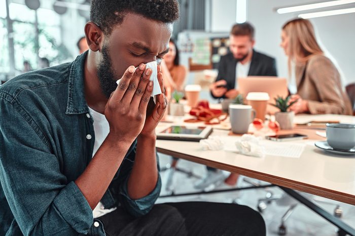 Portrait of ill, sick bearded african male sneezes and coughs, uses handkerchief, rubs nose. Man has running nose, caugh, bad cold, sits at work place