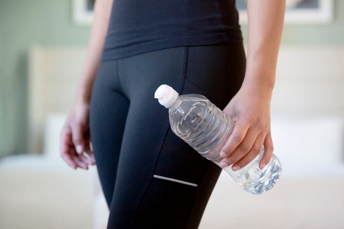 Detail of a mixed race millennial woman's toned midriff, holding a plastic water bottle, wearing black workout pants in her bedroom.