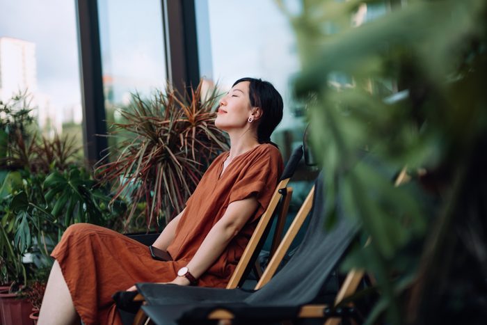 Young Asian woman with eyes closed enjoying fresh air while relaxing on deck chair in balcony in the morning, surrounded by beautiful houseplants. Lifestyle and wellbeing concept