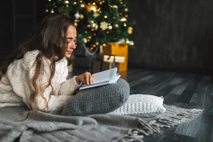 Beautiful woman reading book while relaxing on cushions by Christmas tree at home.