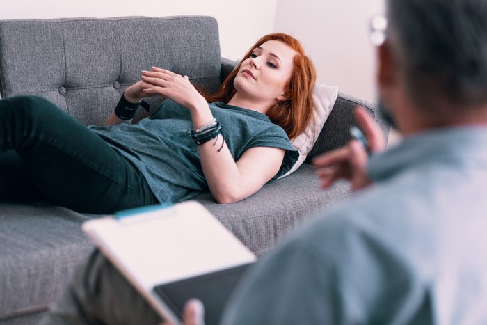Sad young woman lying on a gray couch in psychologist's office. Coping with loss concept. Blurred therapist in the foreground