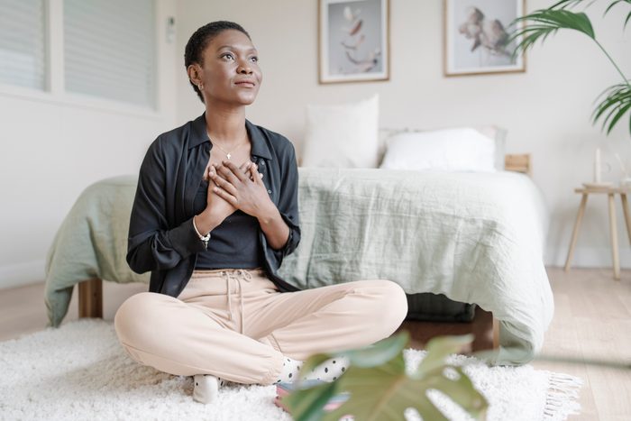 Black woman with hand on chest showing gratitude while praying sitting on the floor in her bedroom.