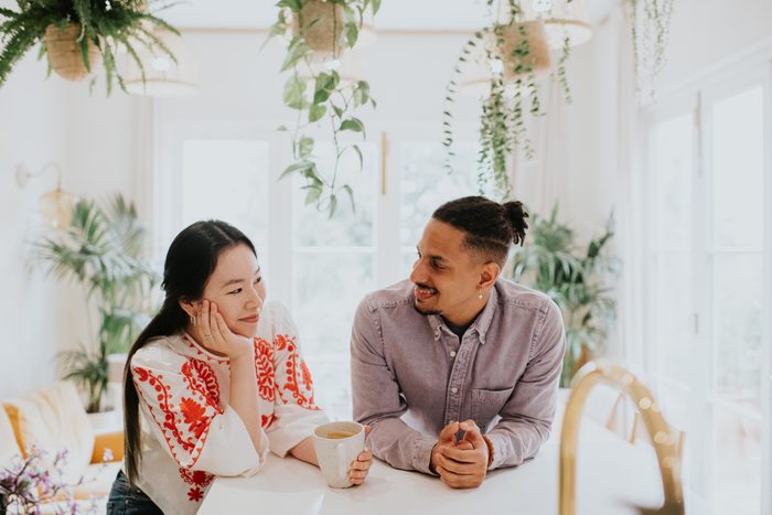 A woman and a man lean on a white surface in a bright, airy room, surrounded by houseplants. They look at each other with fondness and affection.