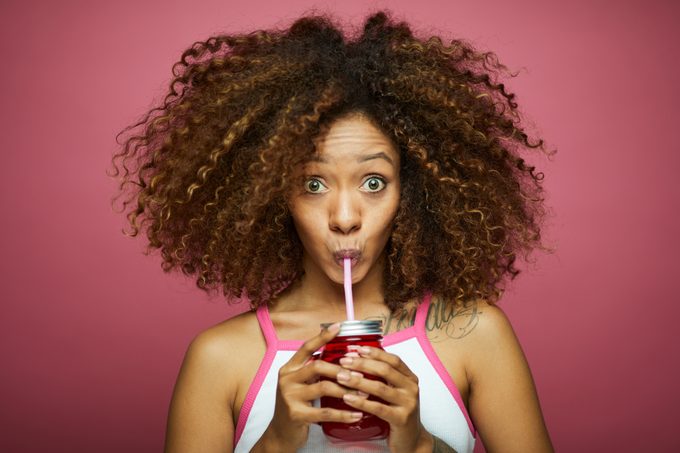 Beautiful afro Caribbean young woman in summer clothes drinks from a straw against pink background