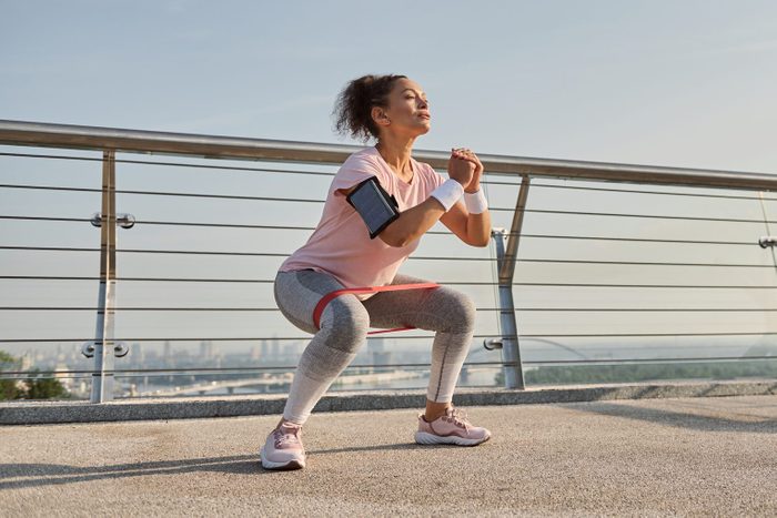 Determined middle aged sporty woman, female athlete exercising outdoor at sunrise, doing squats with elastic resistance band. Fitness sport endurance, body weight training and active lifestyle concept