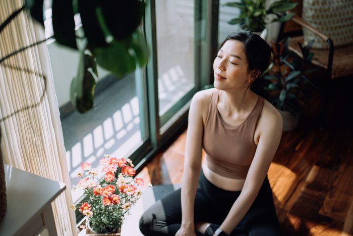 Active young Asian sports woman taking a break after working out at home, sitting on exercise mat taking a deep breath with her eyes closed. Sports and exercise routine. Health, fitness and wellness concept