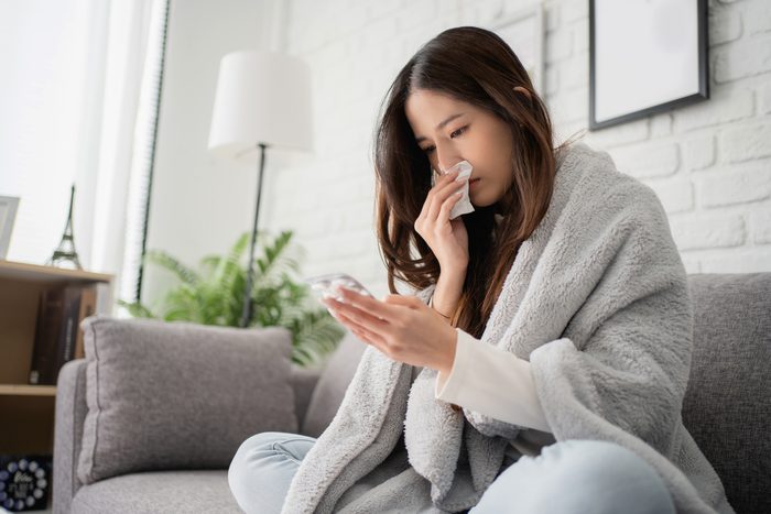 Sick asian woman sit under blanket on sofa and sneeze with tissue paper at home. She is reading check drug medicine capsule at home, suffer from flu. Cold and fever concept.