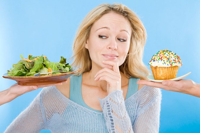 Young Woman Looking At Salad And Cupcake, Hand On Chin, Close Up