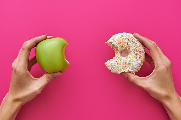 Young Woman Makes Decision of Healthy Versus Unhealthy Foods Holding an Apple and a Cupcake Against a Pink Background