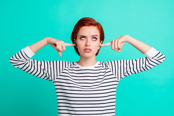 Red Haired lady plugs her ears and rolls her eyes in a striped sweater on a teal background