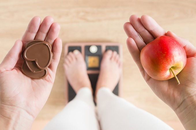 Woman Standing On Scales And Holding Apple And Chocolate Hearts