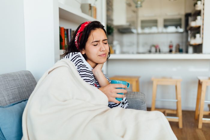 Young Caucasian woman with a sore throat sitting on a couch with blanket and tea