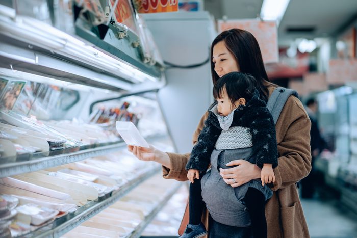 Young Asian mother doing grocery shopping with adorable little daughter. She is choosing for fresh poultry along the refrigerated aisle in a supermarket