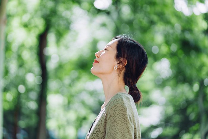 Beautiful young Asian woman meditating in the nature with her eyes closed, setting herself free and feeling relieved. Enjoying fresh air and breathing in the calmness with head up against sunlight in the morning. Freedom in nature. Connection with nature