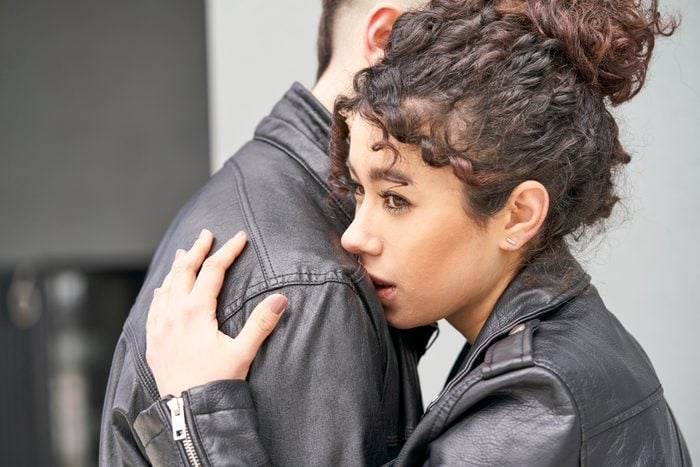 A sad and pensive young woman hugging her boyfriend who is comforting her is standing outside a building.