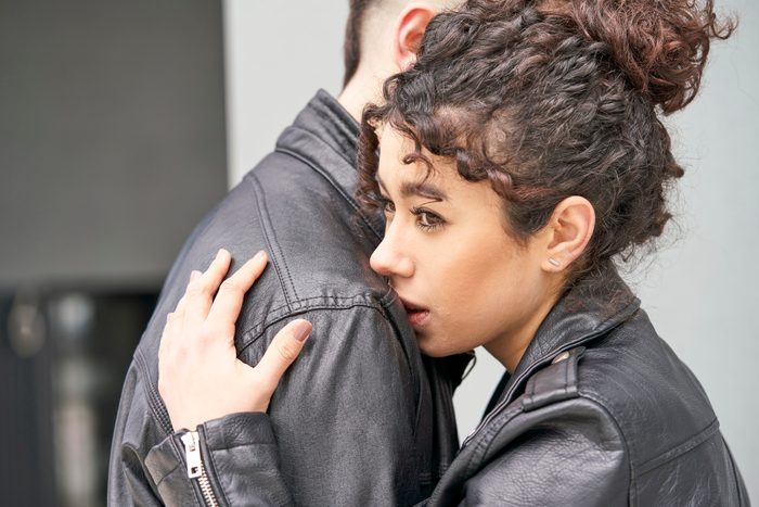 A sad and pensive young woman hugging her boyfriend who is comforting her is standing outside a building.