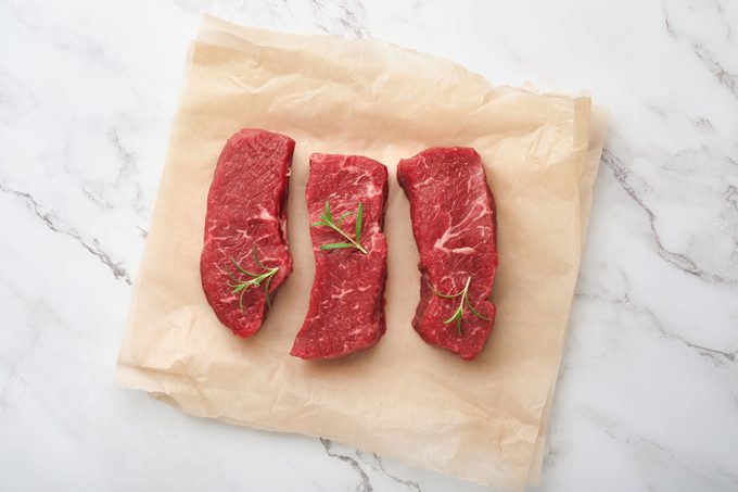 Raw steaks. Top blade steaks on wood burning board with spices, rosemary, vegetables and ingredients for cooking on parchment paper on white background. Top view. Copy space.