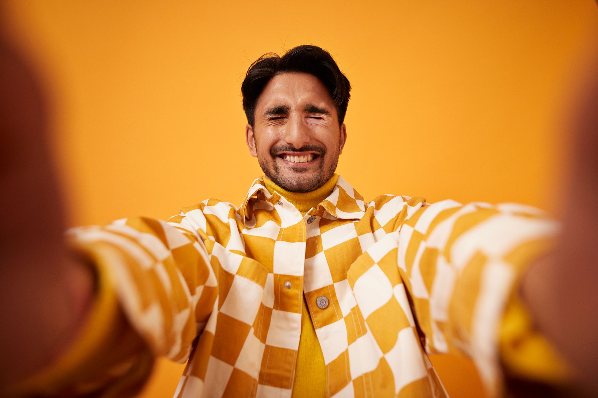 man in a checkered shirt taking a selfie while laughing with eyes closed against dark yellow background