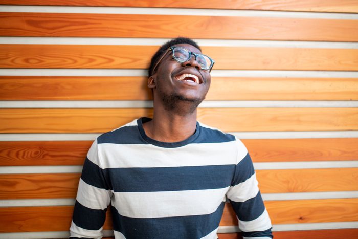 Молодой темнокожий мужчина в полосатой рубашке и очках смеется у стены с полосатым рисунком дерева