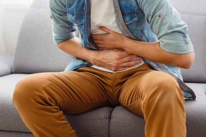 The man is sitting on a gray couch and holding his belly. Medicine and health concept, stomach problems. The man suffers from stomach ache, gastric problems. Abdominal pain, suffering and pain.
