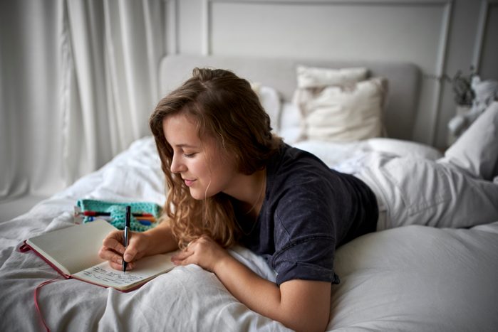 Young woman lying on bed writing in diary for her therapy homework