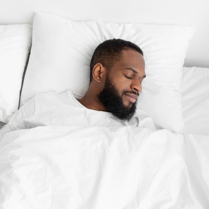 Calm millennial black bearded male sleeping on bed, soft pillow with white duvet in bedroom enjoy rest