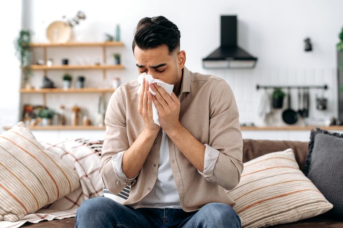 Unhappy sad indian or arabian guy suffering from fever and flu, sitting on sofa in living room, blowing nose and sneezing in napkin, need treatment of illness, and doctors consultation