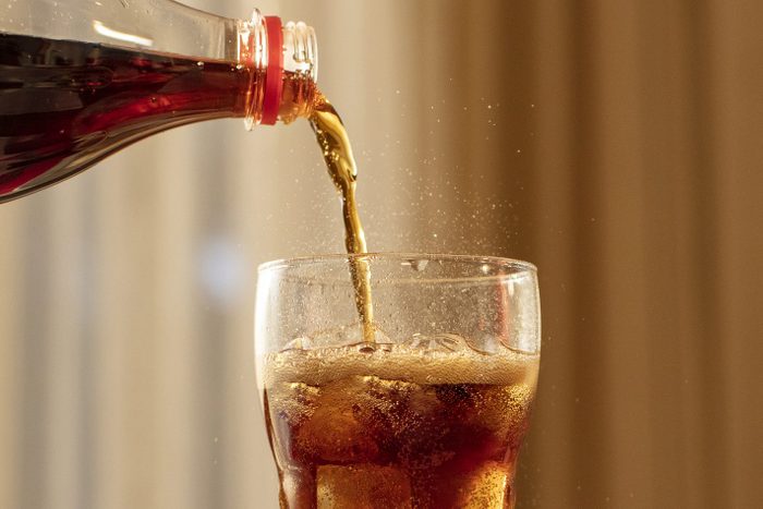 Pouring Cola from Bottle into Glass and Fizz with Ice Cubes on Table Against Blurred Livinroom Background