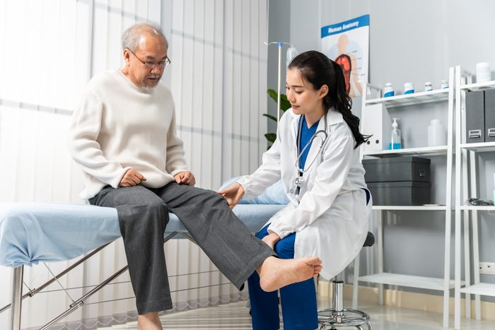 Asian woman physician doing legs raise exercise for senior man patient. Attractive female specialist doctor doing physical therapy procedure for elderly mature male to stretch and recover legs muscle.