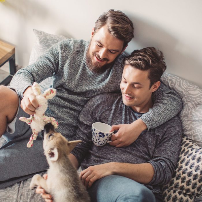 Young gay couple lying on bed in bedroom. Enjoying in morning and first coffee of the day. Wearing pajamas. Caucasian ethnicity, blond hair. Pet dog is with them on bed