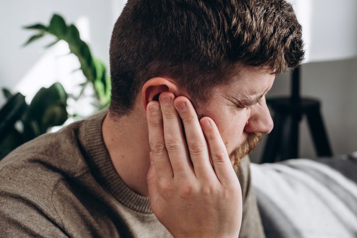 Close-up of unhealthy young caucasian man 30s touching ear, suffering from sudden throbbing ear ache, looking aside. Upset bearded male feeling unwell sitting on couch in living room at home