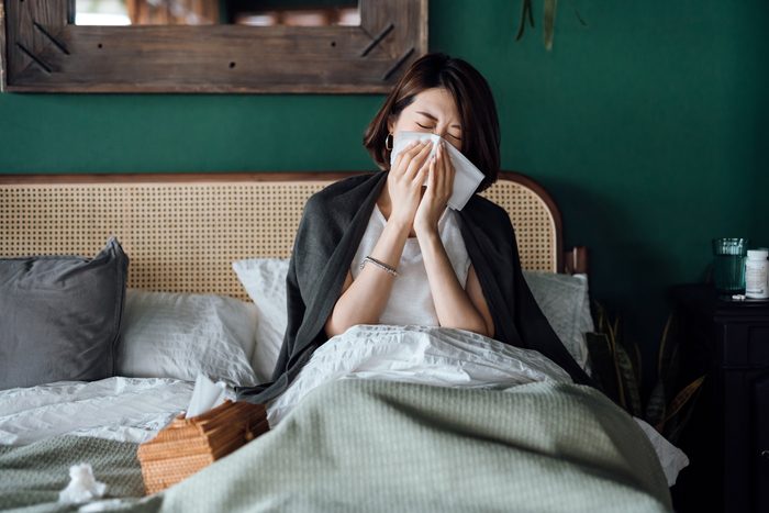 Young Asian woman sitting on bed and blowing her nose with tissue while suffering from a cold, with medicine bottle and a glass of water on the side table