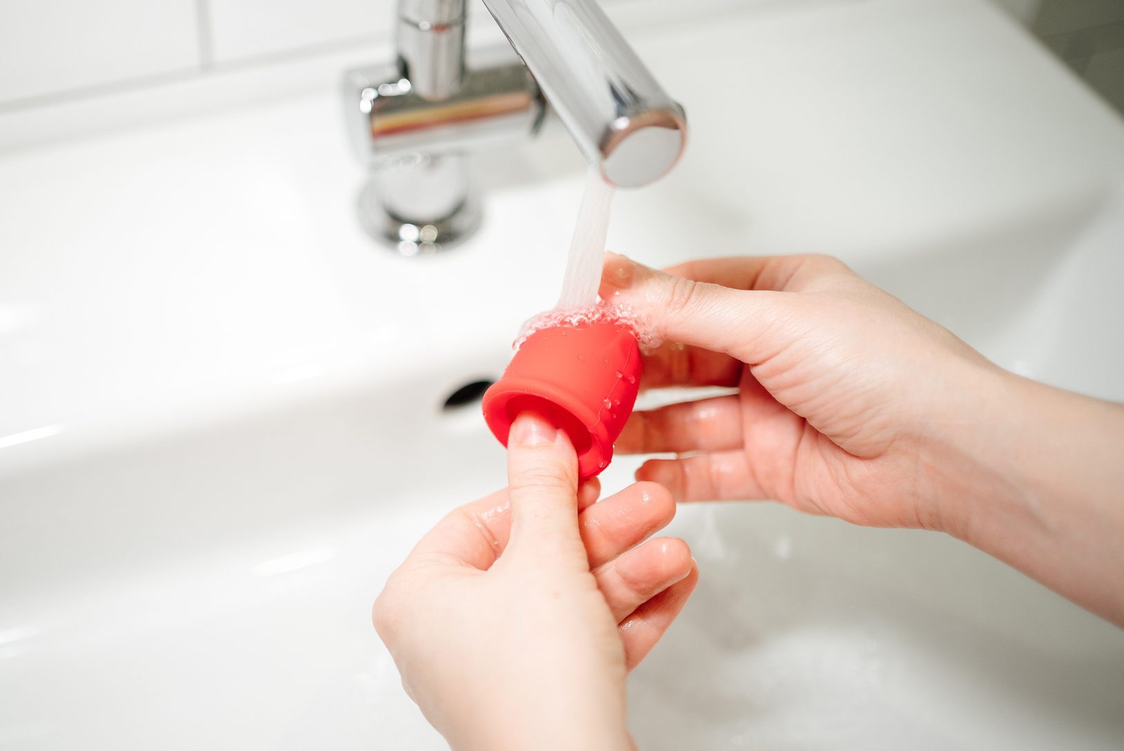 Hands of a woman washes a red menstrual cup with water under the tap at the sink. Alternative feminine hygiene product concept during menstruation.