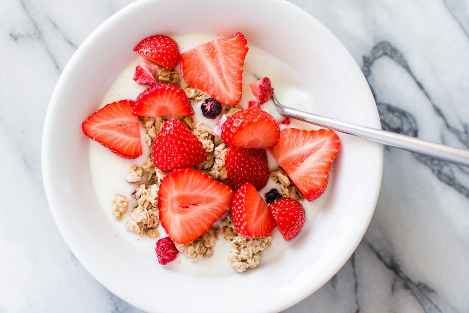 High Angle Overhead View of a White Cereal Bowl with White milk, fresh granola cereal, freshly cut strawberries, on a bright, marble countertop in a residential kitchen