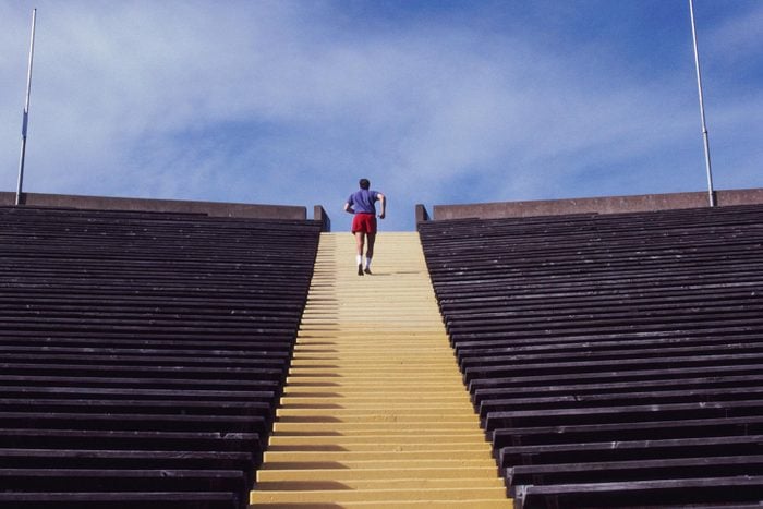 Rear View of Man Running Up Stadium Steps in athletic sneakers, tube socks, a blue t-shirt and red running shorts on a bright, day and blue sky background, no clouds