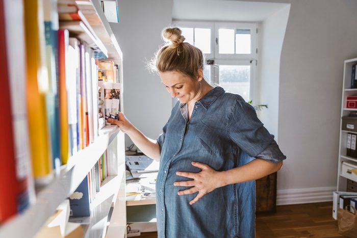 Pregnant woman standing at bookshelf at home, looking down at belly
